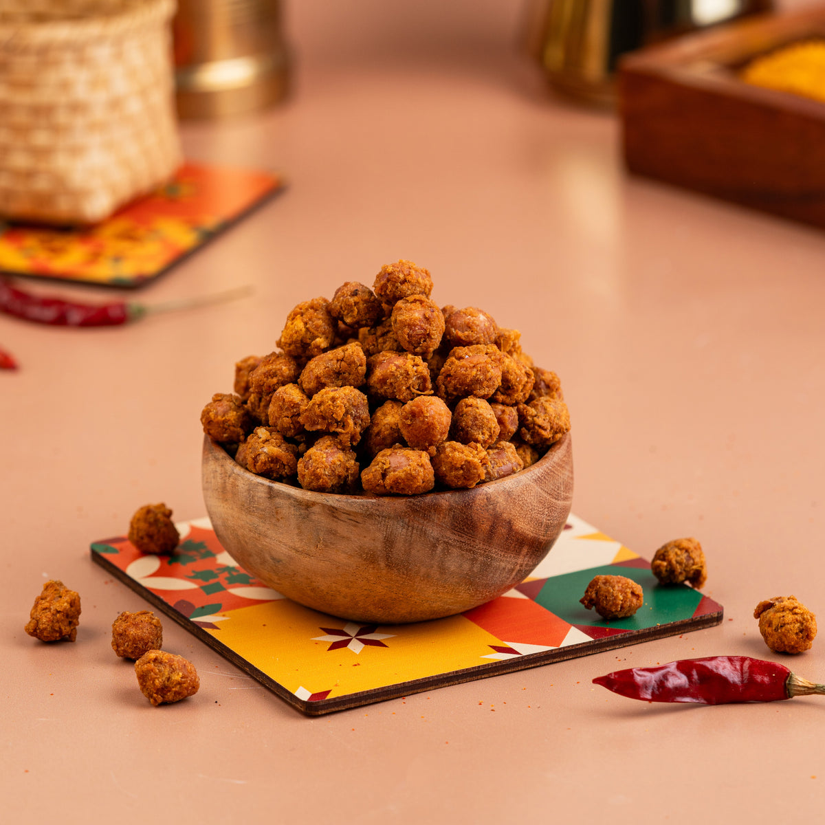 A wooden bowl filled with roasted peanuts, placed on a colorful placemat, with spices and a red chili in the background.