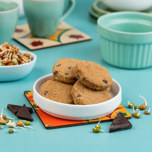 A white bowl filled with chocolate chunk cookies on a colorful mat, with a cup of tea and cereal in the background.