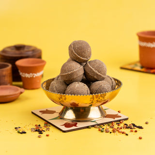 A bowl filled with millet ghee ladoo (Sirudhaniyam) on a yellow background, with pottery and spices scattered around.