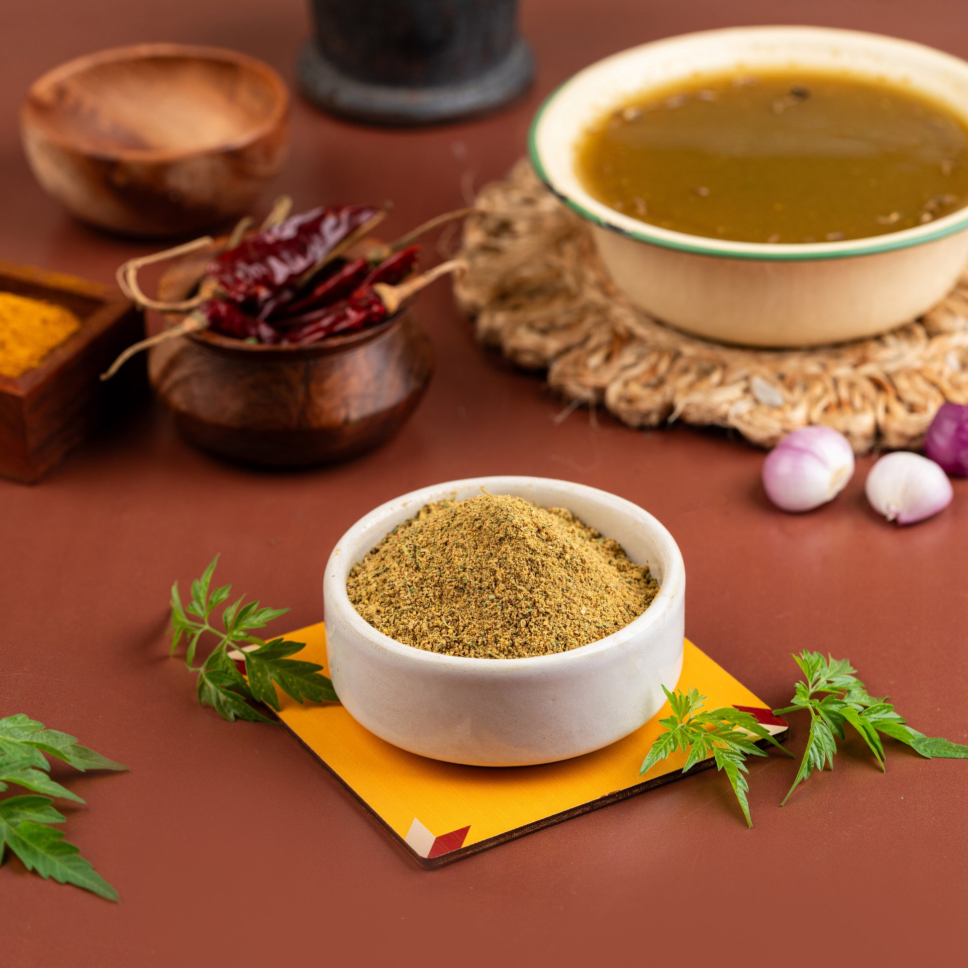 A bowl of Modakthan Soup mix with ingredients such as modakthan leaves, spices, and vegetables around it, presented on a table with a soup bowl in the background.