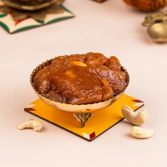 A brass bowl with a golden rim containing a sweet, thick, and syrupy halwa, with peanuts and dried fruits on the side.
