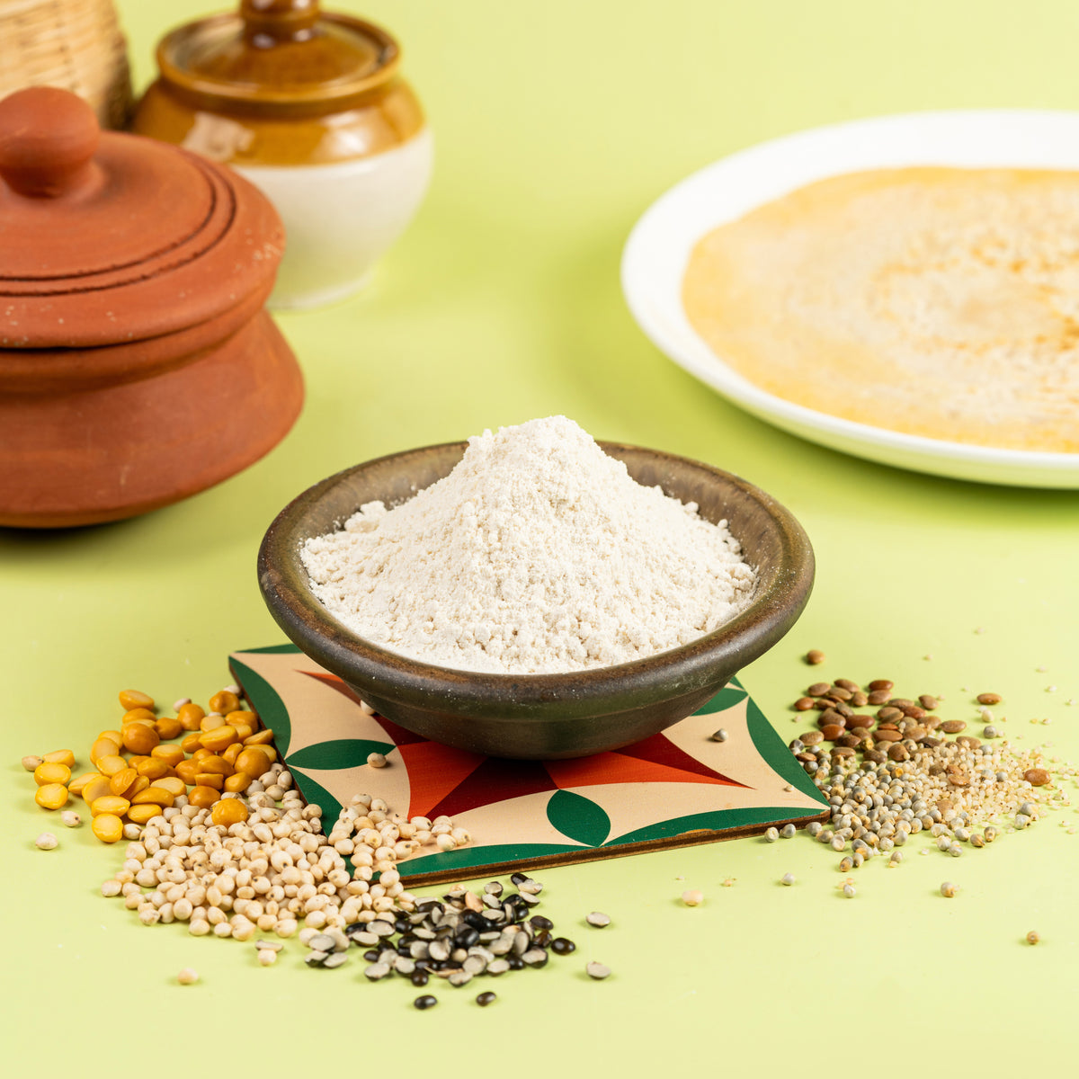 A bowl of Multi Millet Dosa Mix in the foreground with millet grains scattered around, and a dosa (Indian crepe) being cooked in the background on a griddle.