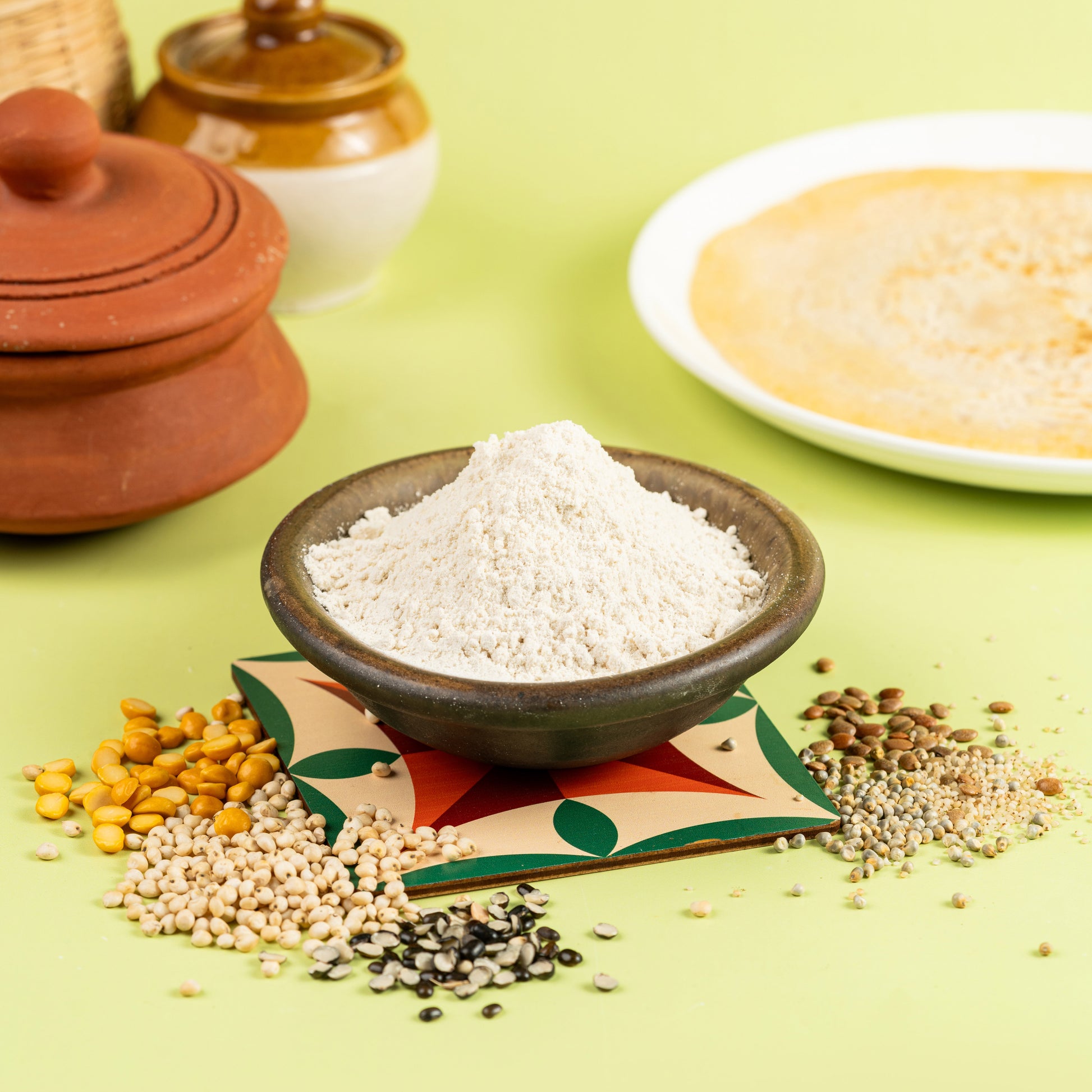 A bowl of Multi Millet Dosa Mix in the foreground with millet grains scattered around, and a dosa (Indian crepe) being cooked in the background on a griddle.