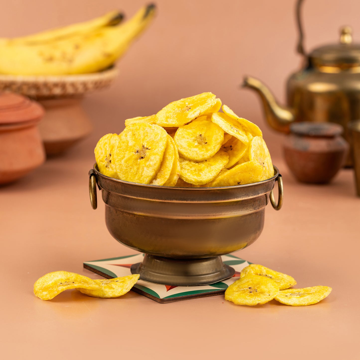 A bowl of crispy banana chips, with a few chips scattered outside the bowl, against a warm-toned background.