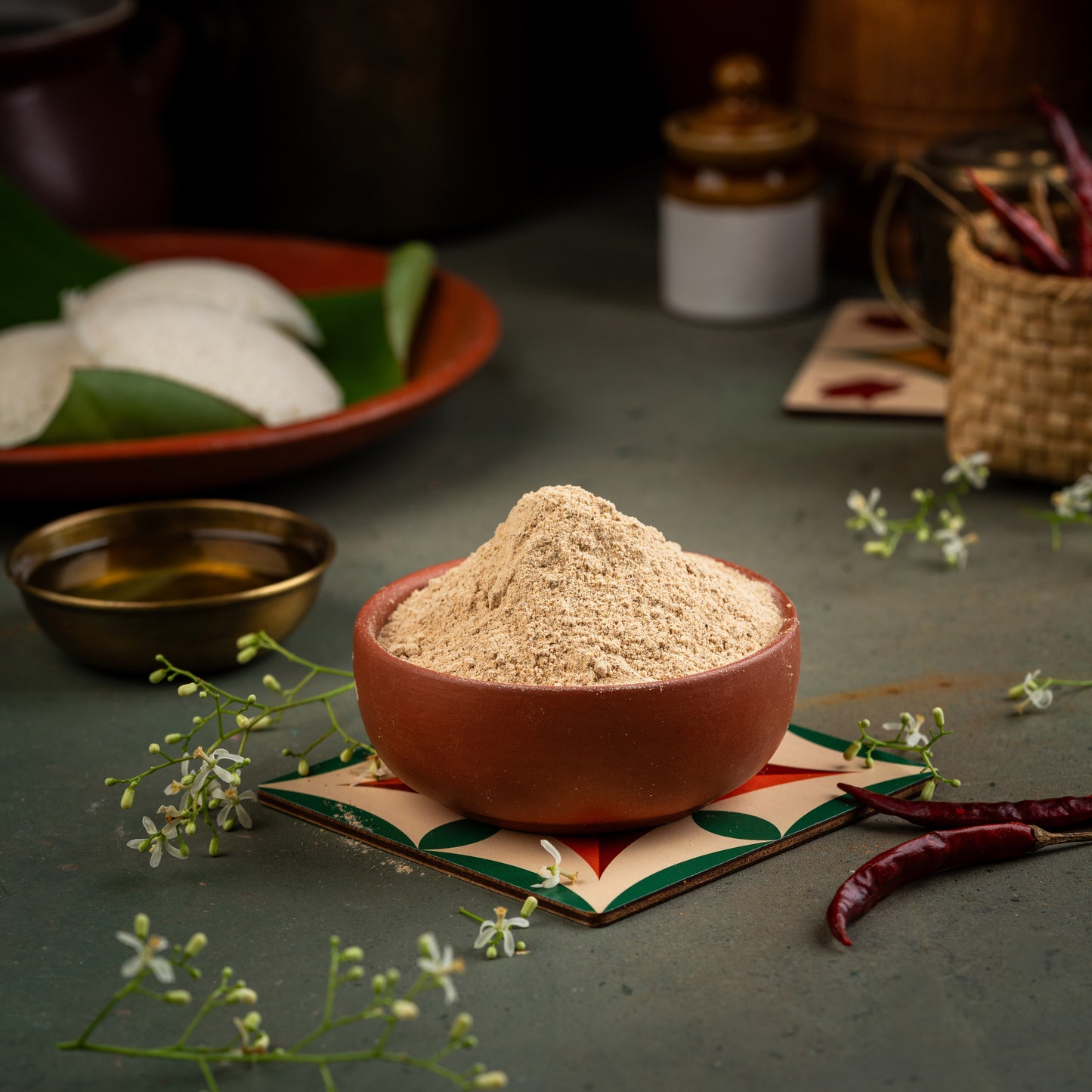 A bowl of Neem Flower Podi (Veppam poo) on a colorful, patterned plate with a small pile of the spice mix, surrounded by various kitchen ingredients like leaves and spices in the background.