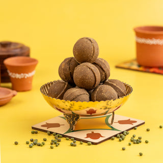 A heap of brown Pasi Payuru (algae lentil) laddoos in a yellow bowl with a ceramic plate, against a yellow background with scattered peas.