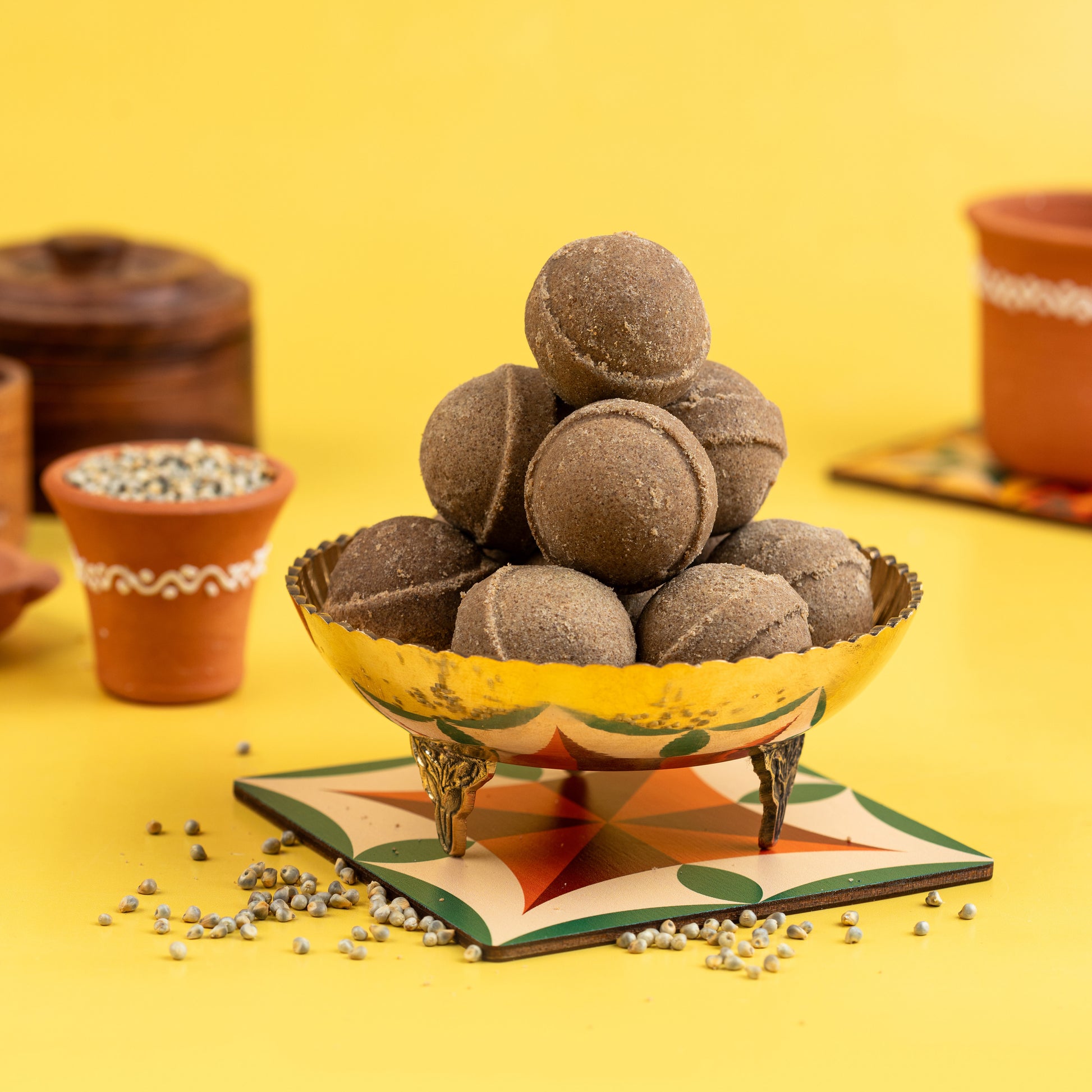 A bowl of pearl millet ghee ladoo (Kambu Ghee Ladoo) served on a decorative plate with scattered millet grains around it, against a yellow background.