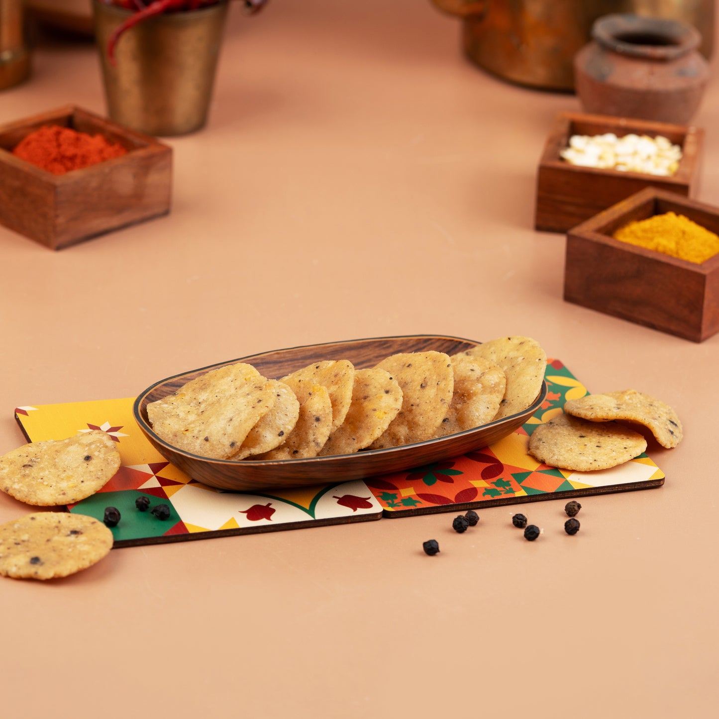A wooden bowl filled with crispy, spicy pepper thattai on a placemat, with spices and condiments in the background.