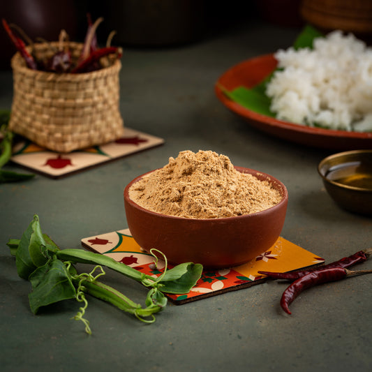 A bowl of Pirandai Podi, a South Indian spice mix, with a small pile of the spice mix in a brown bowl, surrounded by leaves and a piece of ginger on a colorful placemat.
