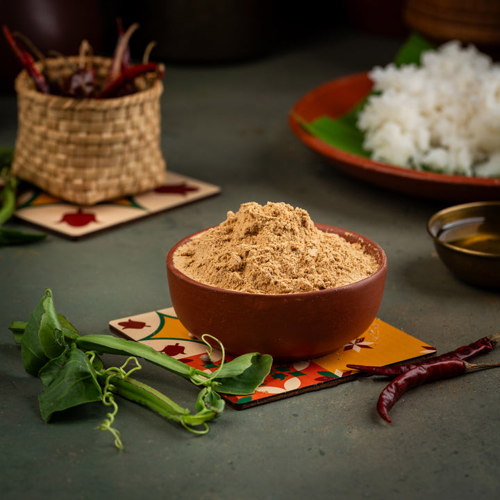 A bowl of Pirandai Podi, a South Indian spice mix, with a small pile of the spice mix in a brown bowl, surrounded by leaves and a piece of ginger on a colorful placemat.