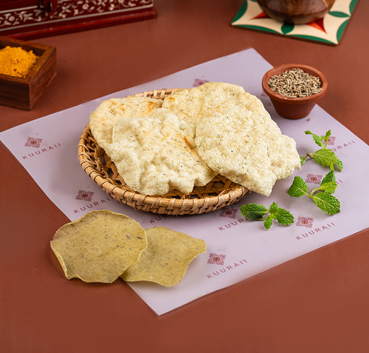 Sun-dried snack foods, possibly a type of vadagam or roasted corn snack, presented on a paper plate with a basket, alongside fresh mint leaves and spices.
