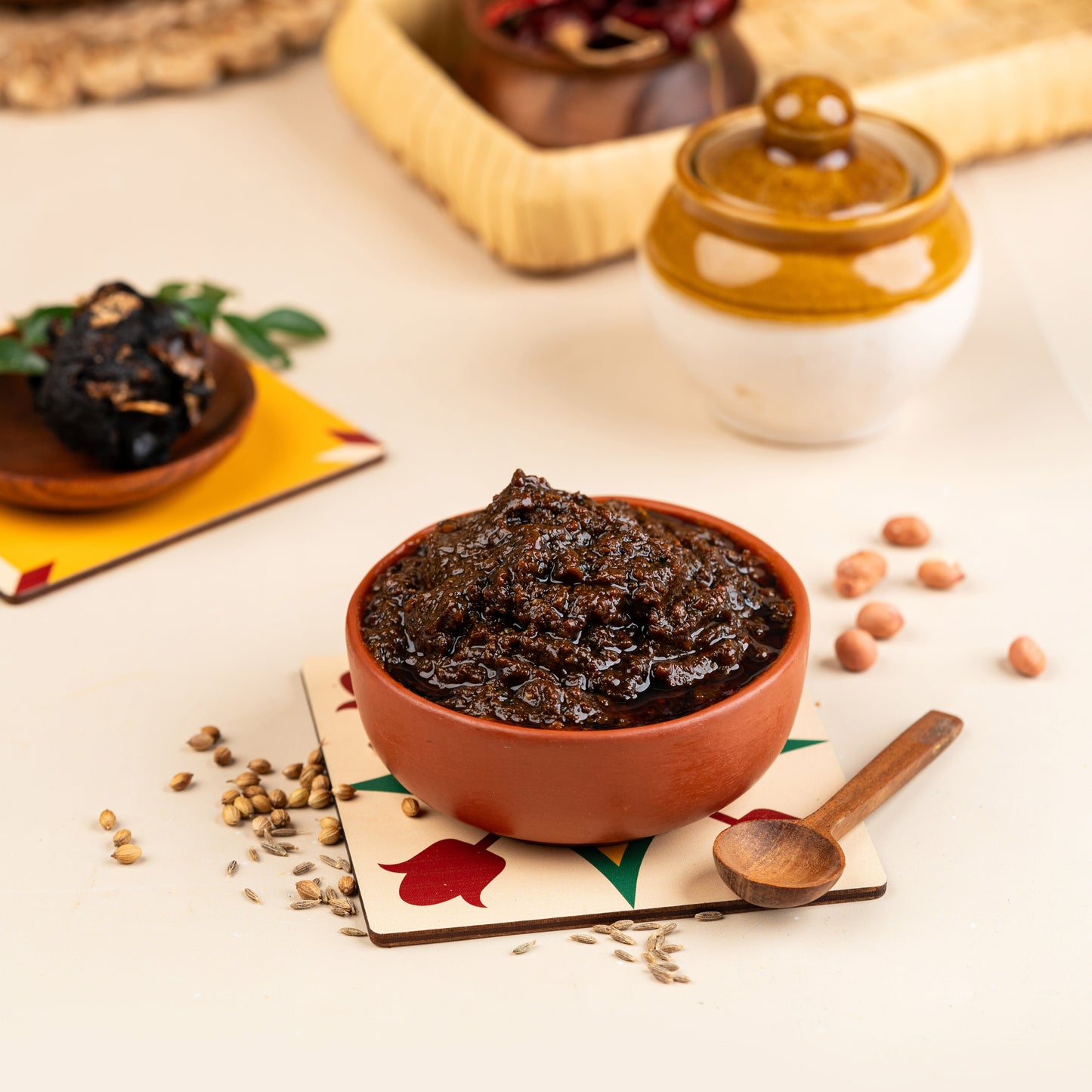 A bowl of Puliyodarai (tamarind rice) mix with spices, alongside a small bowl of tamarind pickle, on a placemat with a spoon resting on it. Background includes other food items and a jar.