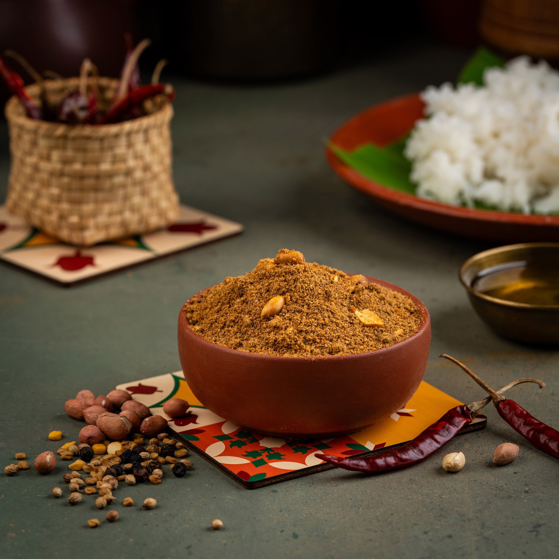 A bowl of Puliyodarai Podi spice mix on a colorful mat, surrounded by spices like mustard seeds, red chili peppers, and spices in the background.