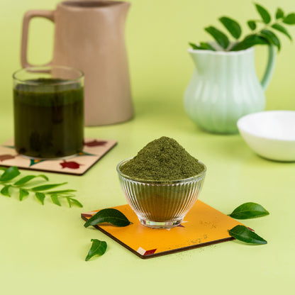 A glass bowl filled with green curry leaf powder, placed on a copper-colored square mat, with a beige background and a green plant in the background.