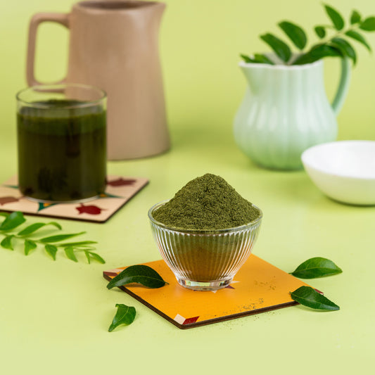 A glass bowl filled with green curry leaf powder, placed on a copper-colored square mat, with a beige background and a green plant in the background.