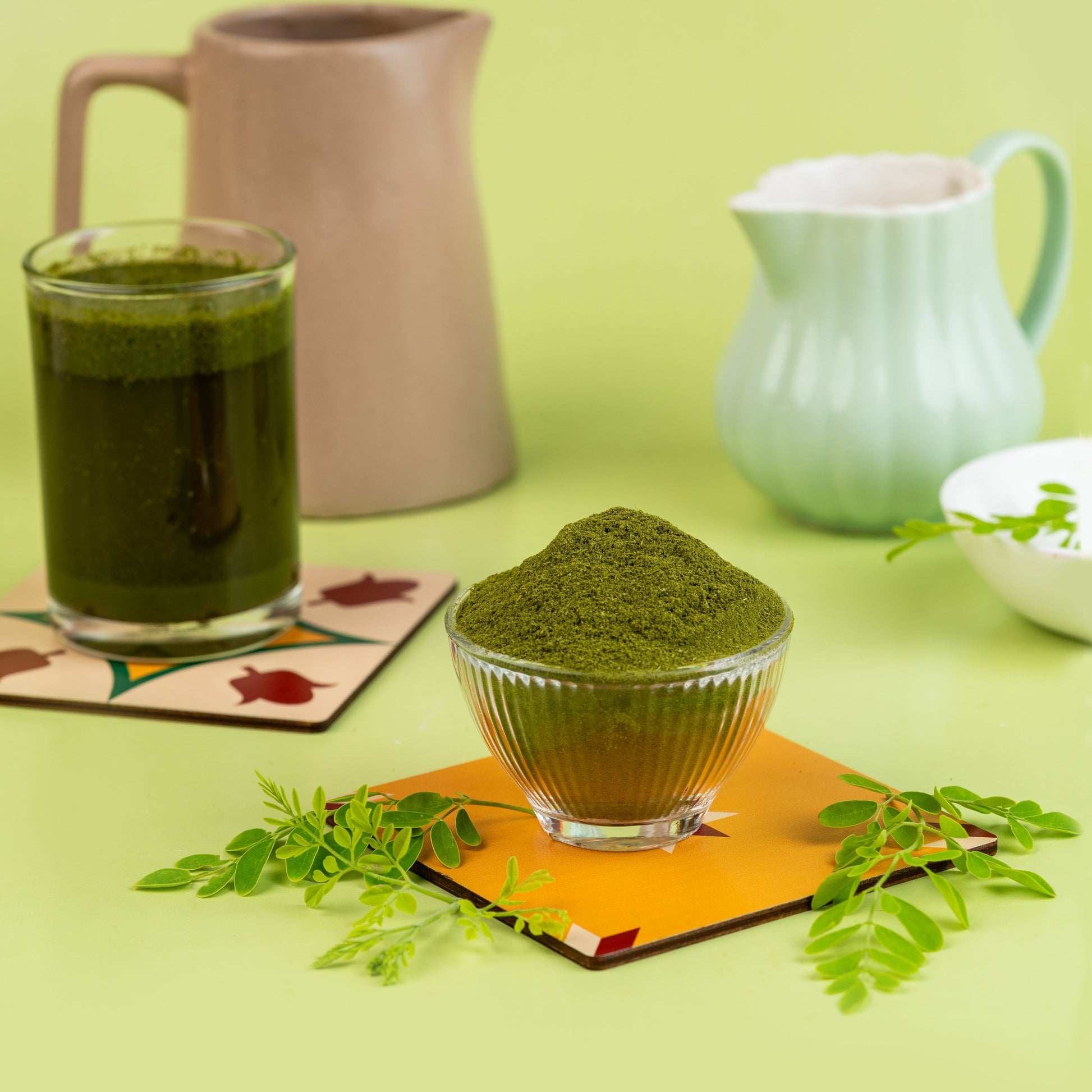 A glass bowl filled with pure Moringa Powder, placed on a coaster with a beige background. There is a cup and a pitcher in the background, and leaves are scattered around the surface.
