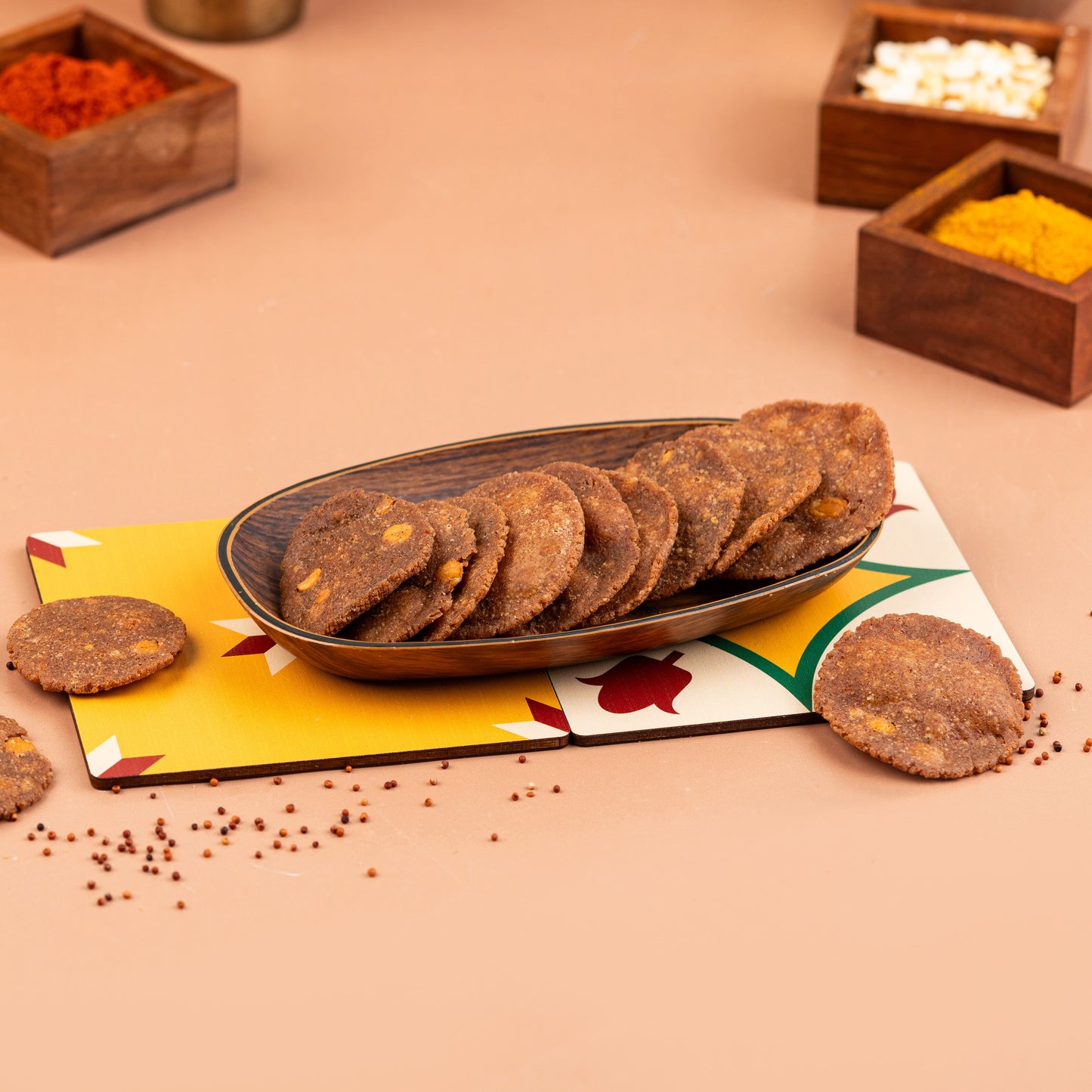 A plate of crispy ragi thattai (finger millet crackers) presented on a colorful mat with wooden containers of spices in the background.