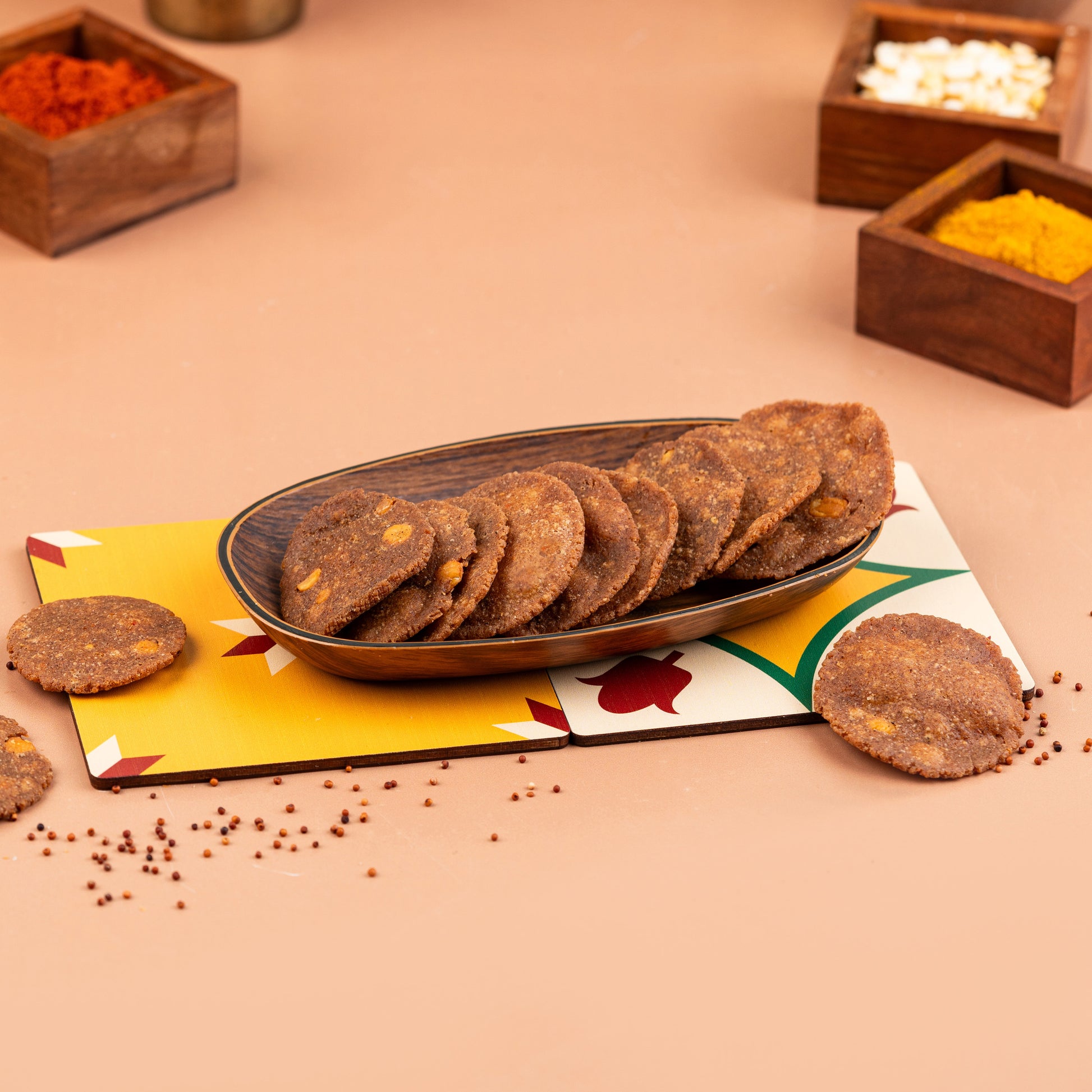 A plate of crispy ragi thattai (finger millet crackers) presented on a colorful mat with wooden containers of spices in the background.