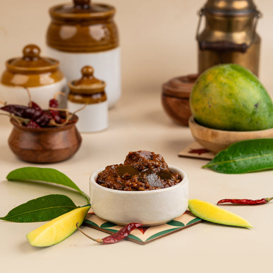 A bowl of raw mango pickle on a mat, with a mango cut in half beside it, and spoonfuls of the pickle, along with whole mangoes and pickle jars in the background.
