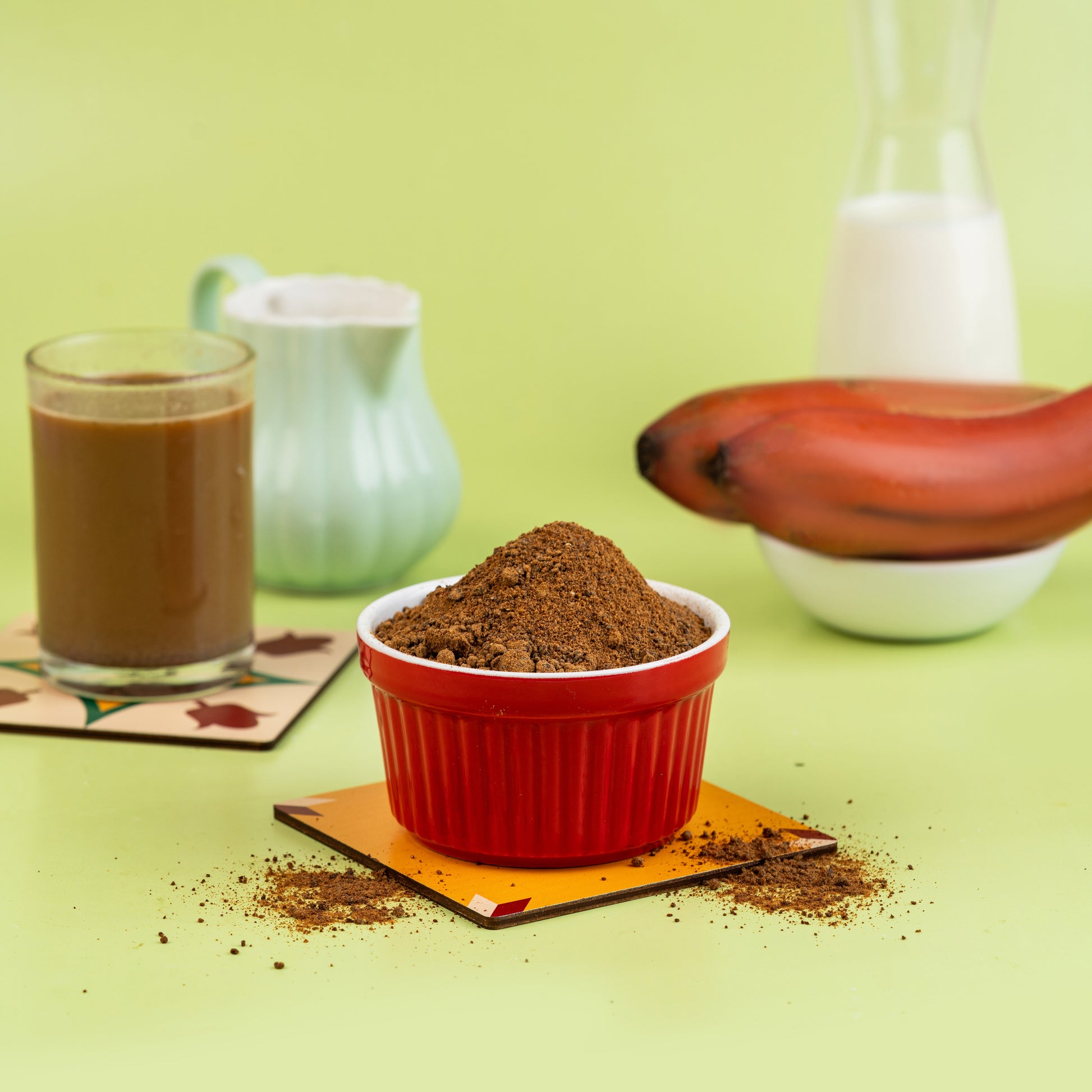 A bowl of Red Banana Malt powder with a glass of milk and a banana in the background, presented on a green surface.