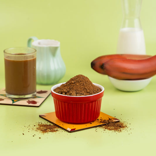 A bowl of Red Banana Malt powder with a glass of milk and a banana in the background, presented on a green surface.