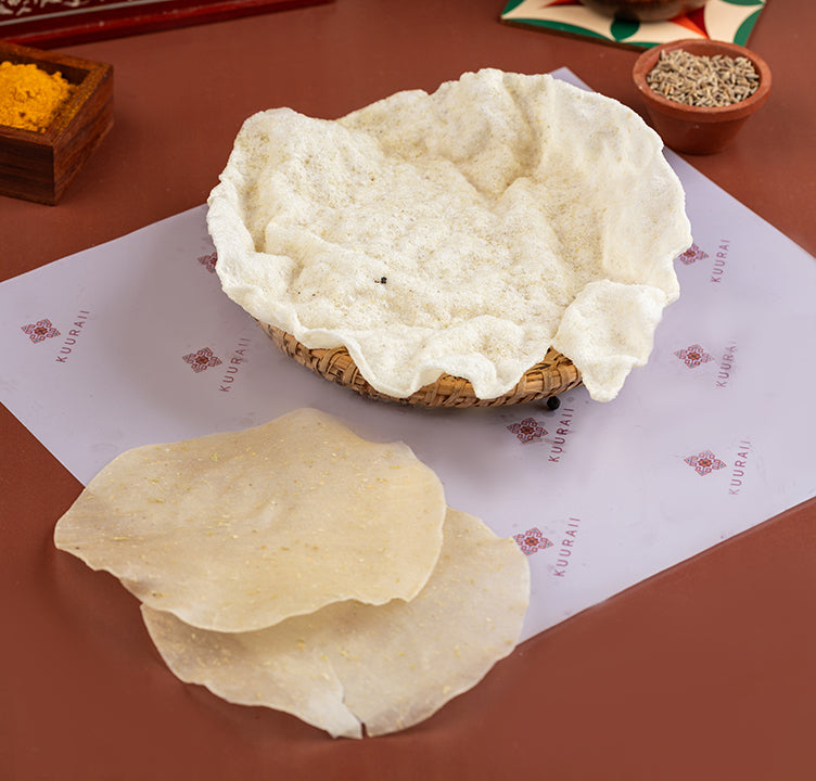 A traditional South Indian crispy wafer, rice appalam, made from rice flour, displayed on a paper. Spices and a serving suggestion are visible in the background.