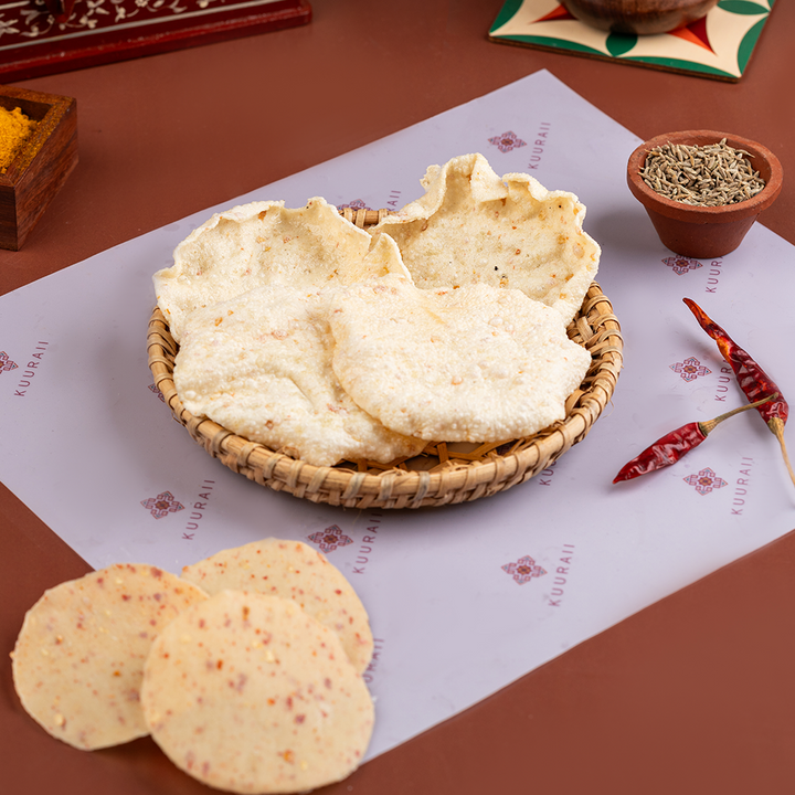 Sun-dried rice cakes with red chili flakes on a flat surface, presented alongside a small bowl of red chili powder and a woven basket.