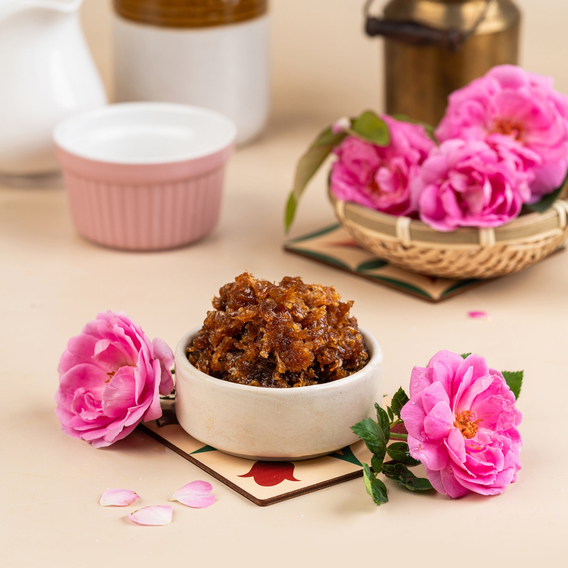 A bowl of Rose Gulkand on a table with pink rose petals, a pot in the background, and a woven basket to the side.