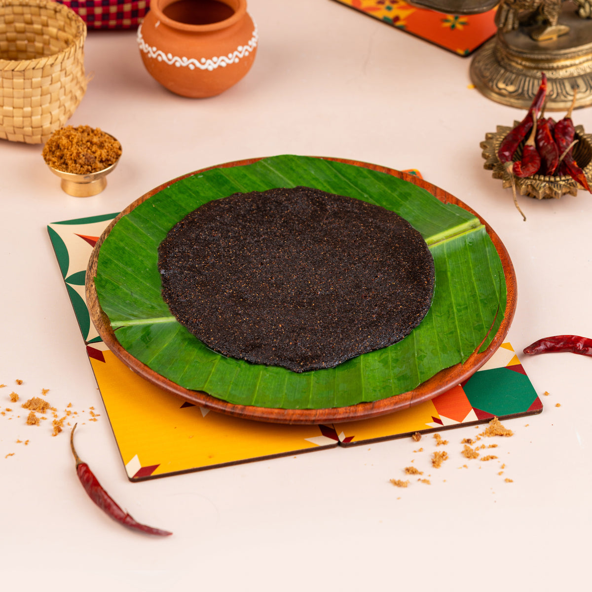 A plate with a green leaf-like border containing a round, dark-colored snack, possibly a Jujube Fritter, served on a table with various colorful items in the background.