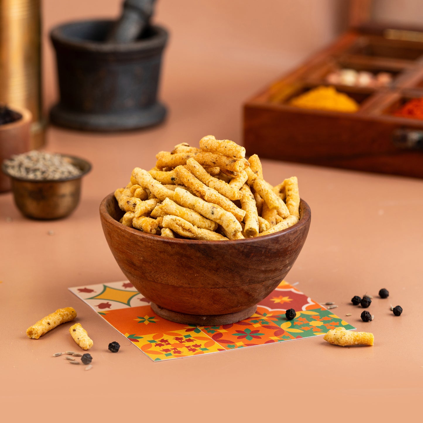 A wooden bowl filled with crispy, peppery rice cakes, presented on a table with spice jars and a colorful fabric underneath.