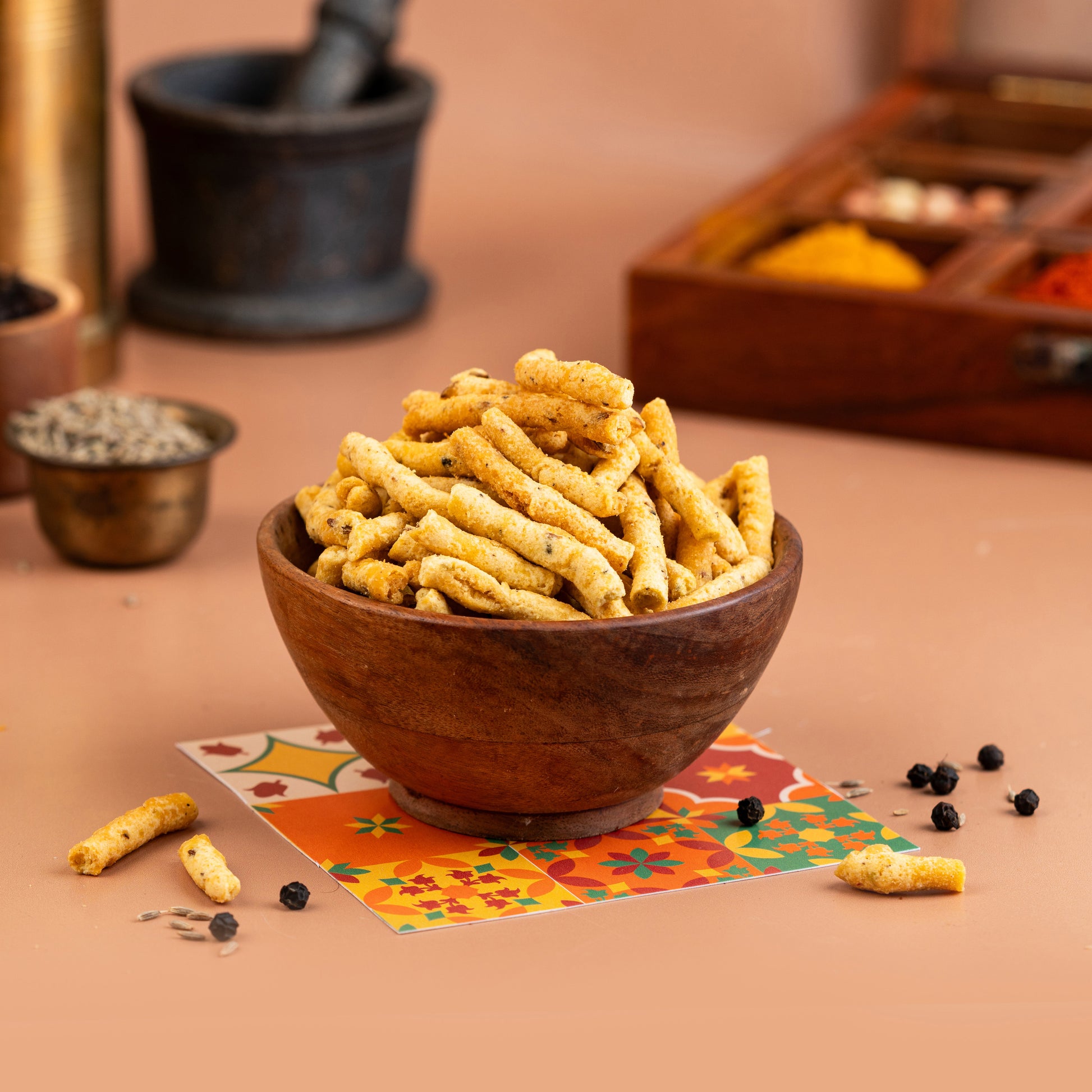 A wooden bowl filled with crispy, peppery rice cakes, presented on a table with spice jars and a colorful fabric underneath.