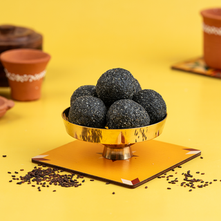 A bunch of black sesame ghee laddooos placed in a gold-colored bowl on a yellow background, with some laddooos scattered outside the bowl.