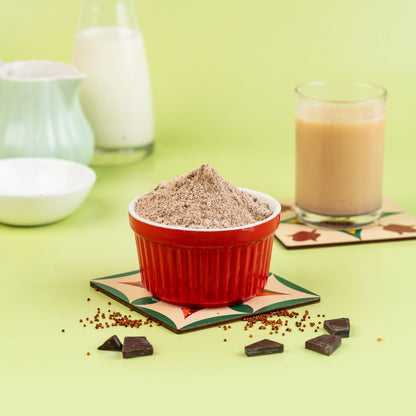 A red bowl filled with sprouted finger millet malt and choco beans on a green mat, with a glass of milk and a bowl in the background.