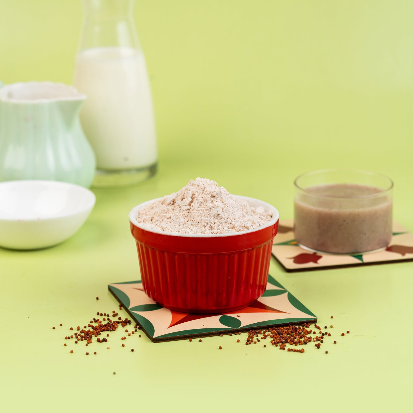 A bowl of sprouted ragi flour is presented in the center with a glass of milk and a cup in the background, all placed on a green surface with patterned coasters.