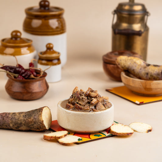 A bowl of Thayir Maganikizhangu Pickle served alongside a slice of sweet potato and pickled vegetables, with traditional ceramic pickle jars in the background.