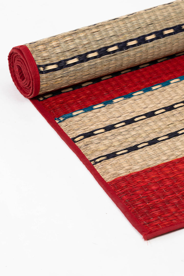 Rolled-up woven grass mat with red and beige pattern on a white background