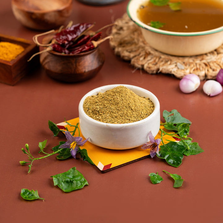 A bowl of Thuthuvalai Soup mix with leafy vegetables, spices, and a touch of herbs, presented on a table with the ingredients and a backdrop of spices.