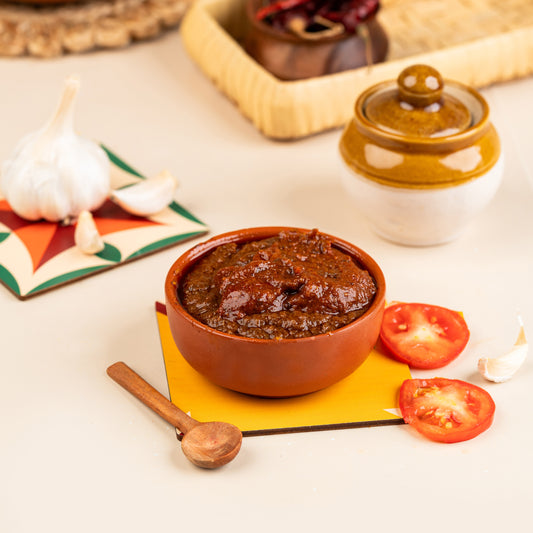 A bowl of tomato and garlic thokku with fresh tomatoes and garlic on a yellow mat, beside a ceramic jar and a wooden spoon.