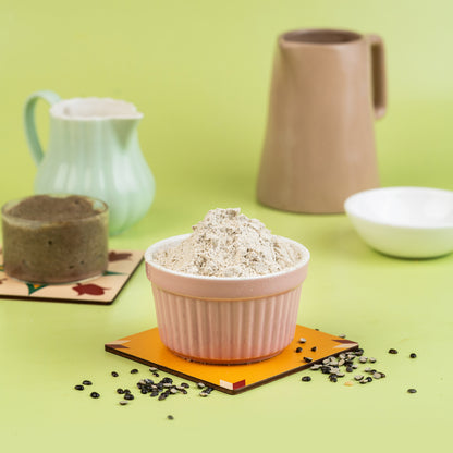 A bowl of Urad Porridge Mix in a pink ceramic cup, surrounded by a green background and other ceramic objects like a mug and a plate.