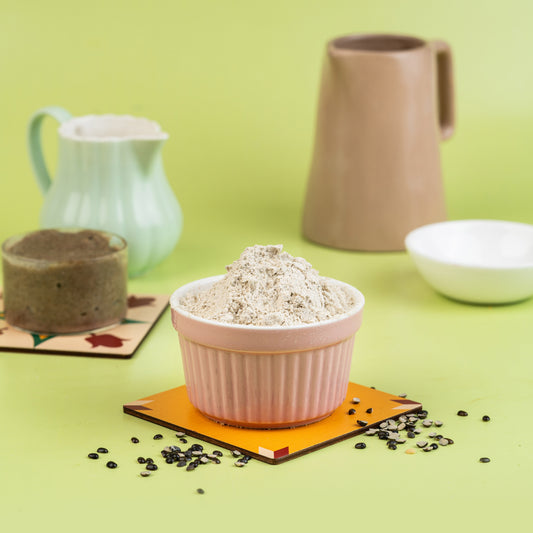 A bowl of Urad Porridge Mix in a pink ceramic cup, surrounded by a green background and other ceramic objects like a mug and a plate.