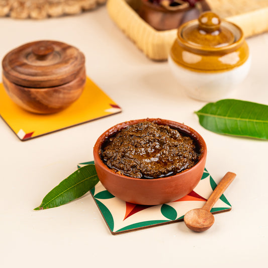 A bowl of Vadumangai Thokku (Baby Raw Mango) served on a colorful napkin with a leaf garnish, accompanied by a wooden spoon and additional cultural decorative items in the background.