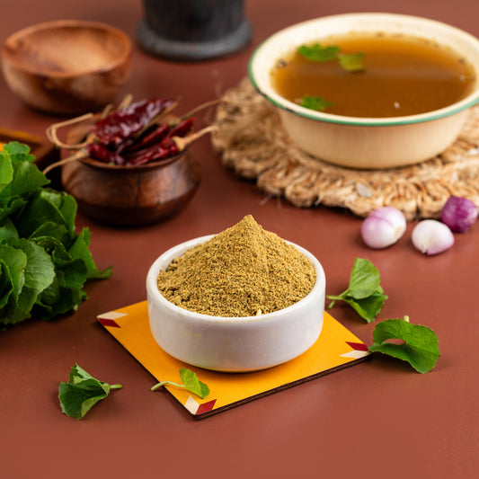 A bowl of Vallarai Soup mix is displayed on a table with a small pile of dried vallarai leaves to the side, and a few whole cloves are also visible. The background includes a spoon, a basket of food, and a pot with more food.