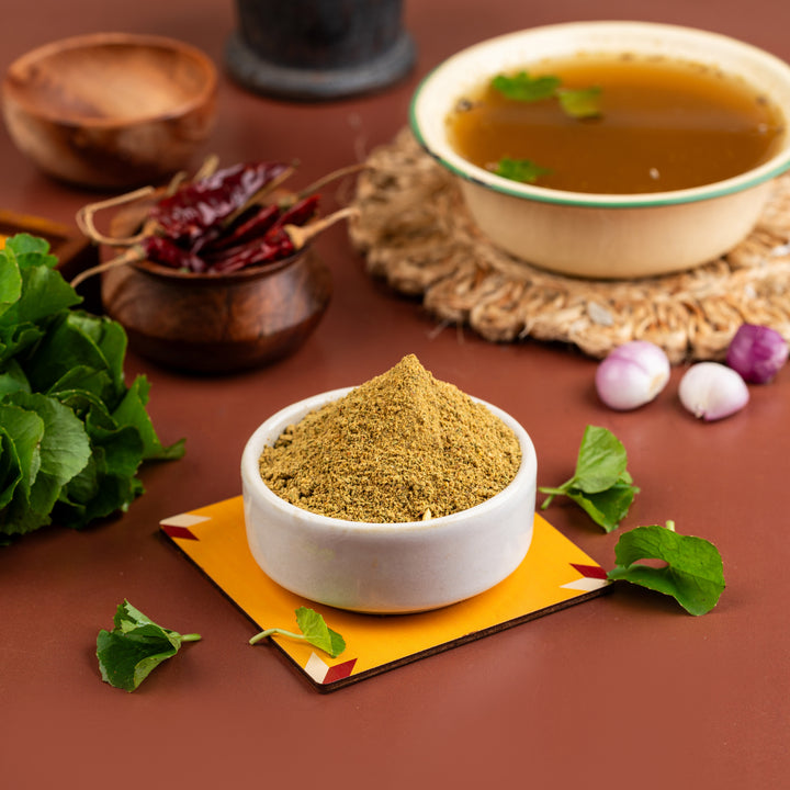 A bowl of Vallarai Soup mix is displayed on a table with a small pile of dried vallarai leaves to the side, and a few whole cloves are also visible. The background includes a spoon, a basket of food, and a pot with more food.
