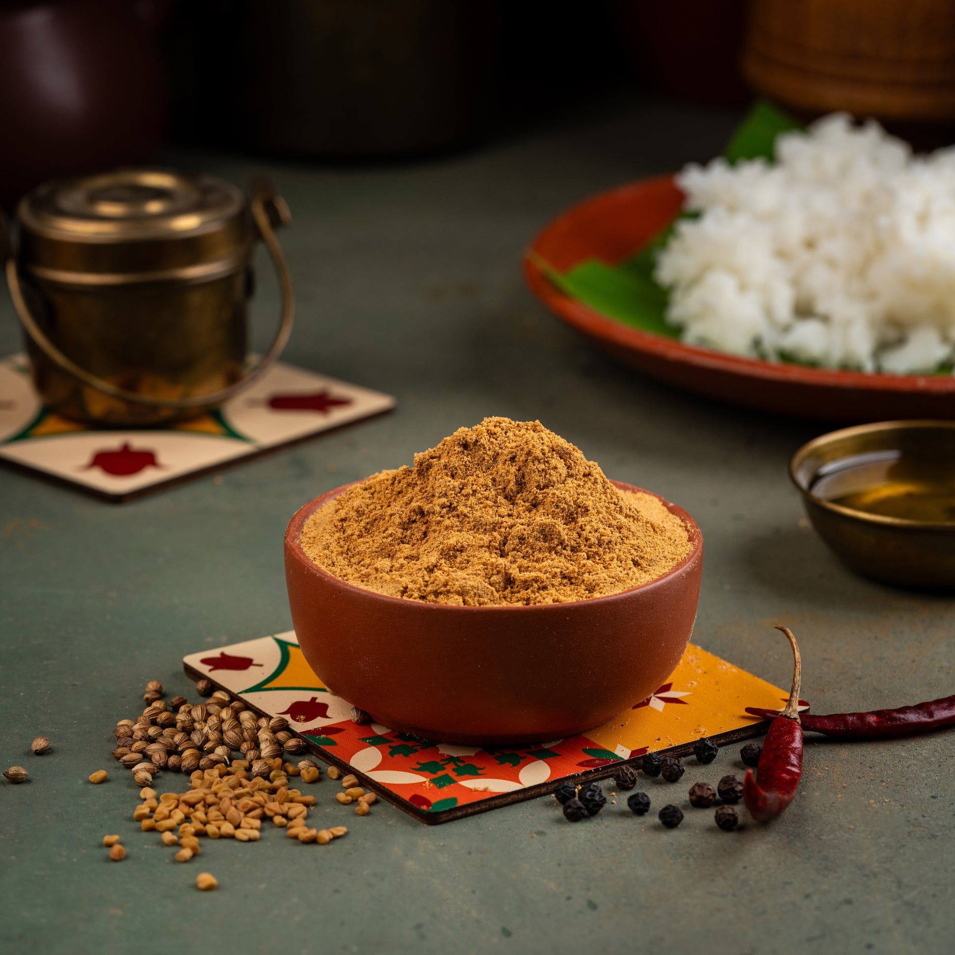 A bowl of Vathakulambu Podi, a South Indian spice mix, with spices like cardamom, cumin, and coriander, displayed on a colorful placemat. In the background, there is a small container of the spice mix and a piece of tamarind.