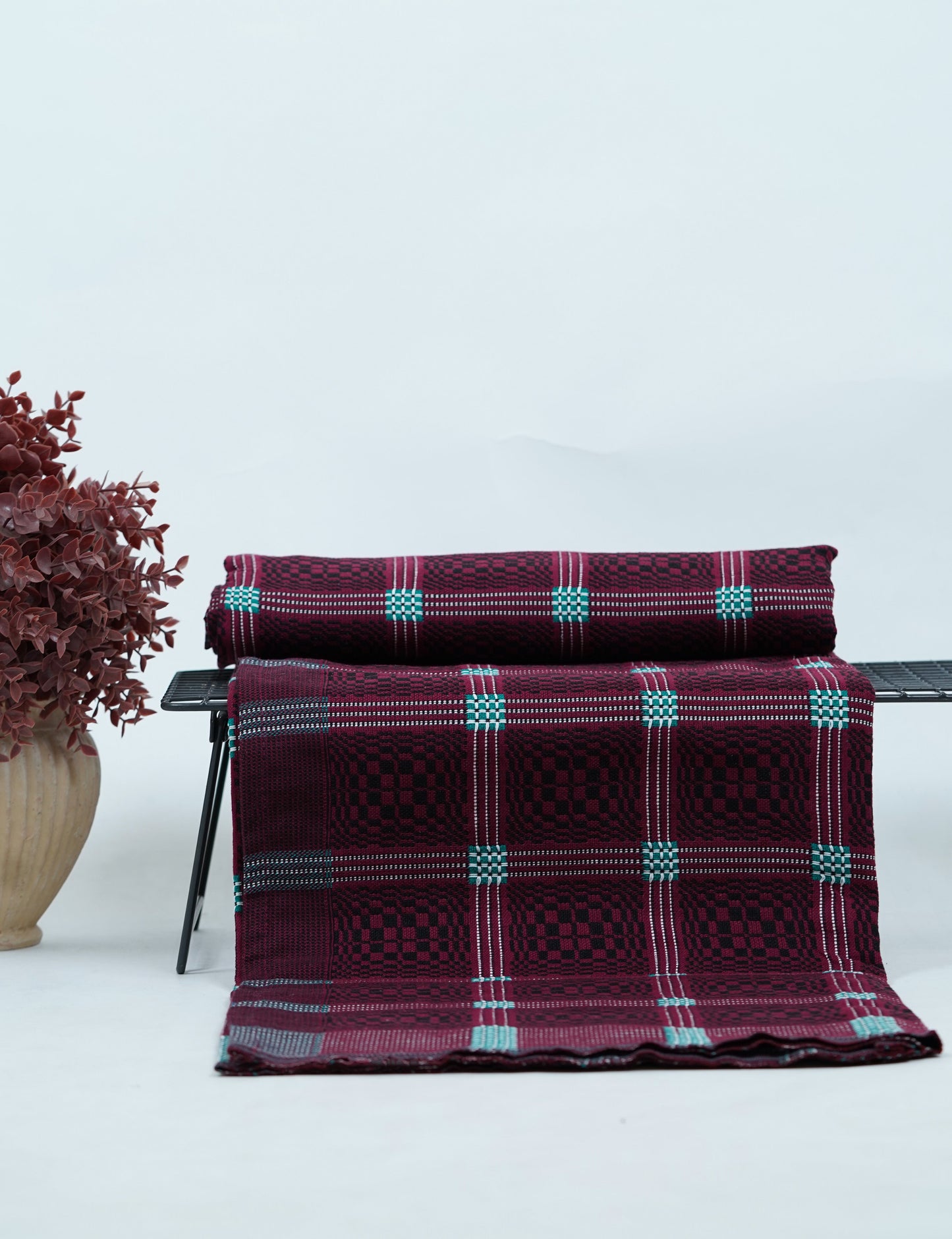 Maroon Nadhiya Chennimalai Bedsheet on a chair with a vase of dried flowers on a light background